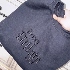 May include: Black sweatshirt with the embroidered text "In my Villain Era" in a gothic font. The sweatshirt is folded and held by a hand, showcasing the design. The garment appears to be made of a soft, dark fabric.