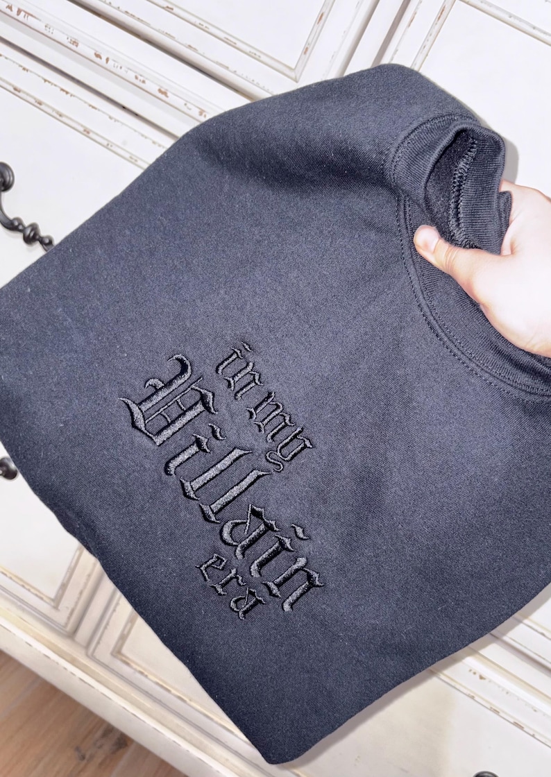 May include: Black sweatshirt with the embroidered text "In my Villain Era" in a gothic font. The sweatshirt is folded and held by a hand, showcasing the design. The garment appears to be made of a soft, dark fabric.