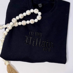 May include: Black crewneck sweatshirt with the phrase "In My Villain Era" embroidered in a gothic font. A decorative string of white and tan wooden beads with a tassel is draped across the sweatshirt.