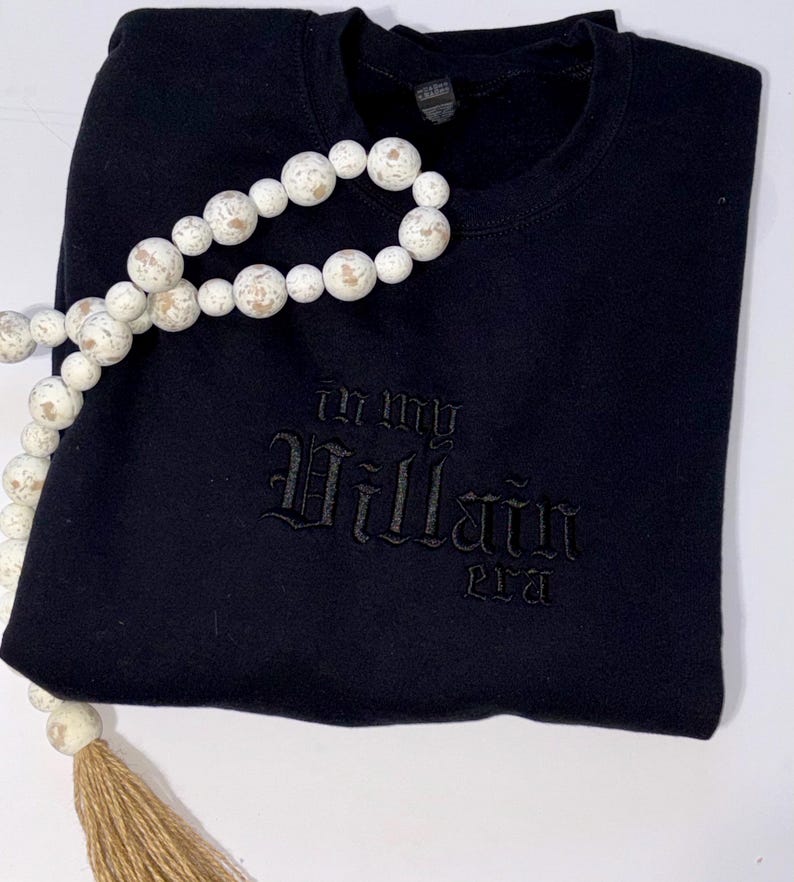 May include: Black crewneck sweatshirt with the phrase "In My Villain Era" embroidered in a gothic font. A decorative string of white and tan wooden beads with a tassel is draped across the sweatshirt.