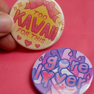 Puede incluir: Dos insignias redondas. Una tiene el texto "Too Kawaii For This" con detalles rosas y un lazo. La otra tiene el texto "Gore Lover" con un diseño de corazón y ojo goteando, en tonos morados y rosas.