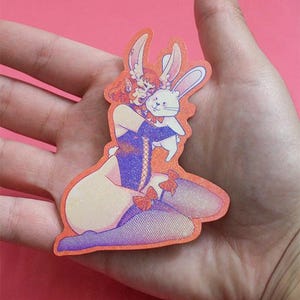 Pode incluir: Um autocolante laranja com uma ilustração de desenho animado de uma pessoa com orelhas de coelho abraçando um coelho branco. A pessoa usa uma roupa roxa com laços vermelhos. O autocolante é segurado em uma mão.