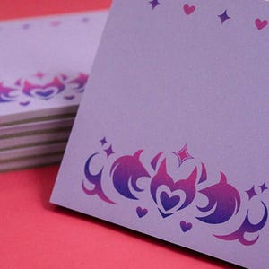 Peut inclure: Une pile de cartes de notes lavande avec une bordure décorative présentant un motif dégradé violet et rose de cœurs, d'étoiles et de formes stylisées en forme de flammes. De petits cœurs et étoiles sont également dispersés en haut des cartes.