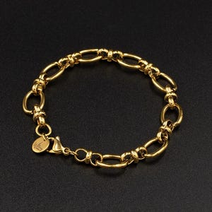 May include: A gold bracelet featuring an oval link chain design. The bracelet has a lobster clasp and a small charm. The jewellery is displayed on a black surface.