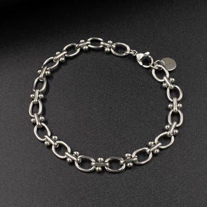 May include: A silver bracelet with a chain-link design. The bracelet features oval links connected by smaller, decorative links with small ball accents. A small, round charm hangs near the clasp.