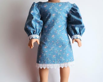 May include: A doll wearing a blue floral print dress with white lace trim and white sandals. The dress has puffy sleeves and a round neckline. The doll has blonde hair and is standing on a wooden surface.