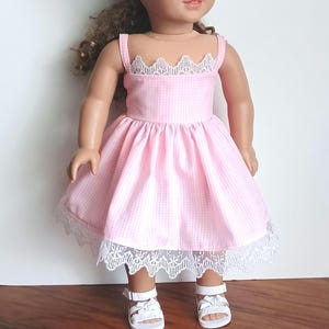 May include: A doll with brown curly hair wearing a light pink and white gingham dress featuring a square neckline with white lace trim and a matching lace hem. The doll is also wearing white strappy sandals with small bow details on the front.