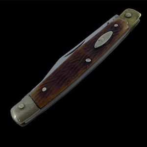 Puede incluir: Una navaja de bolsillo vintage con mango marrón y hoja plateada. El mango tiene un aspecto texturizado con tres remaches plateados y un adorno ovalado decorativo. La navaja está cerrada e inclinada sobre un fondo negro.