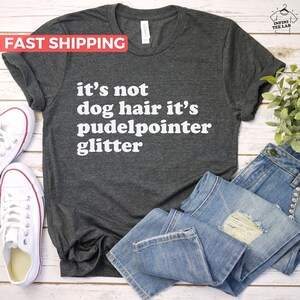 May include: A dark gray t-shirt with white text that reads "it's not dog hair it's pudelpointer glitter". The shirt is laid flat on a wooden surface with a pair of white sneakers and a pair of blue jeans.
