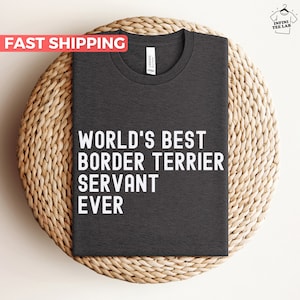Könnte beinhalten: Dunkelgraues T-Shirt mit dem weißen Text "WORLD'S BEST BORDER TERRIER SERVANT EVER". Das T-Shirt wird auf einer gewebten, kreisförmigen Oberfläche präsentiert. Das Shirt hat einen Rundhalsausschnitt und ist aus weichem Material.
