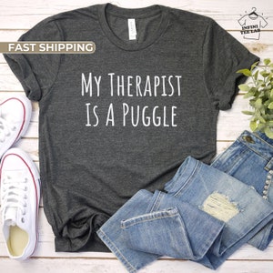 May include: A dark gray t-shirt with the text "My Therapist is a Puggle" printed in white. The shirt is laid flat on a wooden surface with a pair of white sneakers and blue jeans.