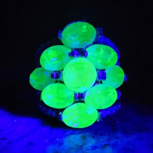 May include: A ring featuring a cluster of bright green, oval-shaped stones set in a silver-toned metal setting. The stones are arranged in a circular pattern, with the largest stone in the centre.