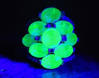 May include: A ring with a cluster of bright green, oval-shaped stones set in a silver-toned metal setting. The stones are arranged in a circular pattern, with the largest stone in the center.
