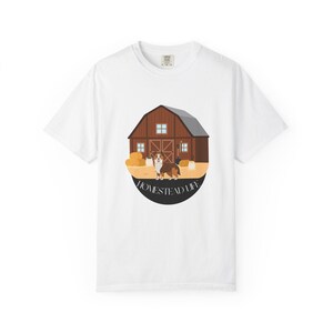 Puede incluir: Una camiseta blanca de manga corta con un gráfico circular de un granero marrón con pacas de heno y un perro delante. El texto "HOMESTEAD LIFE" está impreso en un arco negro en la parte inferior del diseño circular en el centro.