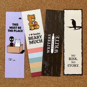 May include: Four colorful bookmarks with unique designs. One features an alien and cat in a box with the text "THIS MUST BE THE PLACE." Another has a bear with the text "I ❤️ books BEARY MUCH." A third says "WRITERS GONNA WRITE." The last shows a raven and says "NO RISK. NO STORY."