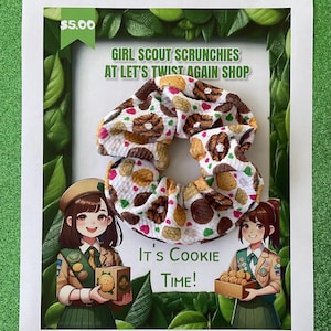 May include: A white scrunchie with a pattern of Girl Scout cookies, including chocolate and peanut butter cookies. The image also features the text "Girl Scout Scrunchies at Let's Twist Again Shop" and "It's Cookie Time!" with illustrations of Girl Scouts.