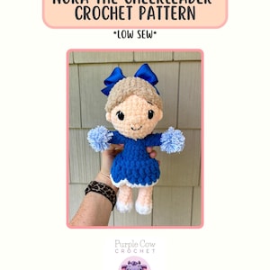 May include: A crocheted cheerleader doll with a blue dress, light brown hair, and blue bows. The text "NORA THE CHEERLEADER CROCHET PATTERN" is displayed above the doll. The text "*LOW SEW*" is also displayed. The doll is handmade.