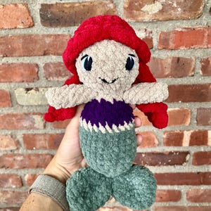 May include: A crocheted plush toy mermaid with a red hat, purple top, and green tail. The mermaid has blue eyes and a smiling face.