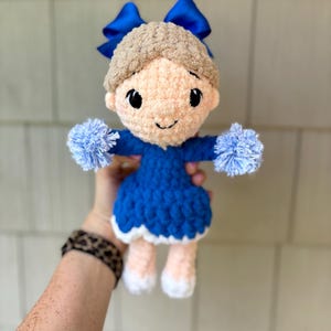 May include: A handmade crochet doll with a blue dress, blue pom-poms, and a blue bow. The doll has light brown hair and a smiling face. The doll's legs are peach-colored with white feet.