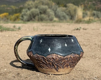 May include: A ceramic mug with a dark blue upper half and a brown, textured mountain design on the lower half. The mug has a handle and white star accents. The mug is sitting on sand.