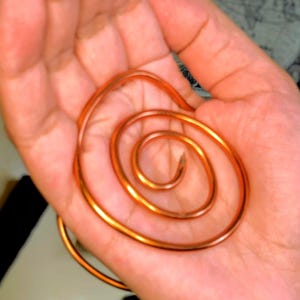 May include: A copper wire spiral, held in a hand. The spiral is a warm, metallic orange colour, with a smooth, reflective surface. The wire is coiled tightly, forming a central point and expanding outwards. The background is a blurred view of the hand.