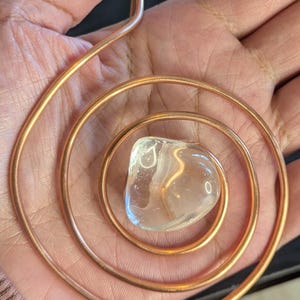 Op de afbeelding: Een koperdraad hanger in de vorm van een platte spiraal met een enkele heldere, gepolijste, onregelmatig gevormde steen in het midden. De metalen draad heeft een warme, roodbruine tint en een gladde, afgeronde afwerking in een spiraal.