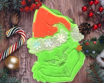 May include: Green Grinch character with a red Santa hat and white snowflakes on a wooden background. The Grinch is made of a soft, green material and has a grumpy expression.