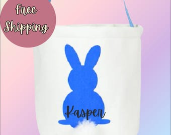 Puede incluir: Una cesta de Pascua de lona blanca con una silueta de conejo azul y el nombre "Kasper" impreso debajo. El conejo tiene una cola blanca y esponjosa.