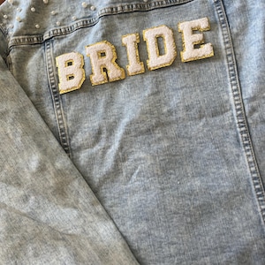 May include: Light blue denim jacket with the word "BRIDE" in white, gold-trimmed letters across the back. The jacket is embellished with small, scattered pearl beads. A stylish bridal jacket.