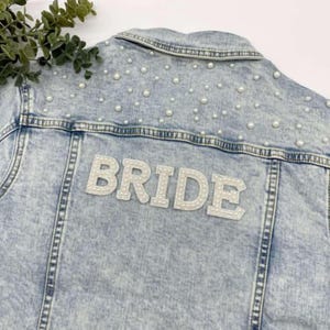 Könnte beinhalten: Hellblaue Jeansjacke mit Perlenbesatz und dem Wort "BRIDE" in weißen, perlenbesetzten Buchstaben auf dem Rücken. Die Jacke hat einen klassischen Kragen und eine Knopfleiste.