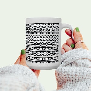 May include: White ceramic mug with a repeating black geometric pattern. The mug has a curved handle. The design consists of various shapes and lines arranged in horizontal bands around the mug. The mug is held by a person.