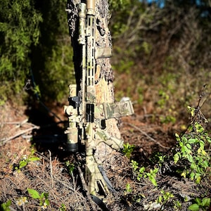 Puede incluir: Un rifle con estampado de camuflaje, con mira telescópica y silenciador, se apoya en un tronco de árbol en una zona boscosa.