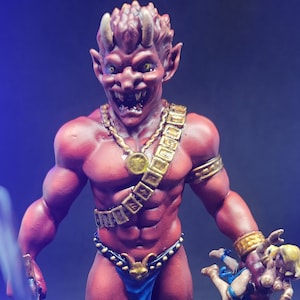 May include: A red, horned, and muscular figurine holding a smaller, female figurine. The larger figurine is wearing a gold necklace, a gold belt, and a blue loincloth. The smaller figurine is wearing a blue and red dress. Both figurines are painted in detail.