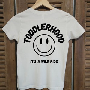 Könnte beinhalten: Weißes T-Shirt mit schwarzem Text "Toddlerhood It's a wild ride" und einem schwarzen Smiley.