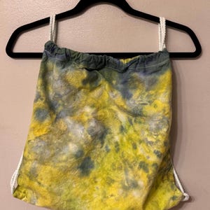May include: A yellow and blue tie-dye drawstring bag with white drawstrings. The bag is hanging on a black hanger.