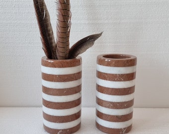 May include: Two cylindrical vases with horizontal stripes of brown and white marble. One vase contains several brown and tan feathers. The vases are of similar size and shape, and are suitable for home decor.
