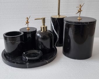 Può includere: Un set da bagno in marmo nero con dettagli dorati. Il set comprende un dosatore di sapone, un bicchiere, un vassoio e un contenitore coperto. Ogni pezzo è decorato con una piccola statuina di ballerina dorata.