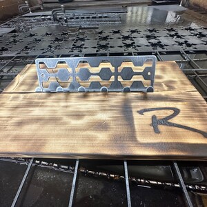 May include: A metal key rack with a geometric design, mounted on a wooden board with a burnt finish. The board has the letter "R" burned into it.