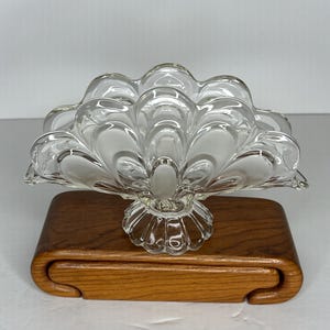 May include: A clear glass decorative dish with a fan-like design, resting on a wooden base. The dish features a scalloped edge and a pedestal base. The wooden base has a curved design. The dish is likely for decorative purposes.