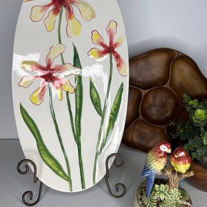 May include: A white oval ceramic serving platter with a floral design featuring yellow, red, and green flowers. The platter is on a black metal stand. A ceramic figurine of two colorful parrots perched on a branch is in the foreground.