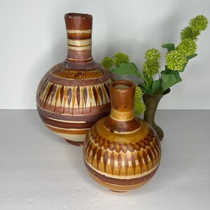May include: Two handcrafted, brown and beige ceramic vases with decorative patterns. The larger vase has a rounded body and a long neck, while the smaller vase is rounder. Green floral accents are in the background.