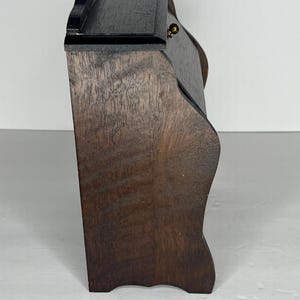 Puede incluir: Una caja de madera marr&oacute;n oscuro con una tapa con bisagras y un peque&ntilde;o pomo de lat&oacute;n. La caja tiene un frente ligeramente curvado y un borde festoneado en la parte superior.