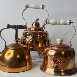 May include: Three copper tea kettles with different designs. The kettles have curved spouts, lids, and handles. One kettle has a wooden handle, while the others have white ceramic handles. The kettles are displayed against a white background.