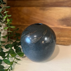 May include: A small, round, dark blue ceramic vase with a silver leaf design. The vase is sitting on a white surface.