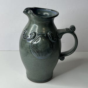 May include: A handcrafted, ceramic pitcher in a muted teal color. The pitcher features a seashell design and a curved handle. The rim and the seashell design have a subtle blue accent. The pitcher is suitable for serving beverages.