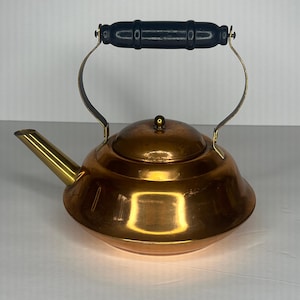 May include: A copper teapot with a black wooden handle and a brass spout. The teapot has a rounded body with a slightly flared lip.