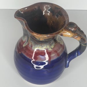 May include: A small, blue and brown ceramic pitcher with a unique handle. The pitcher has a glazed finish and a slightly rounded shape.