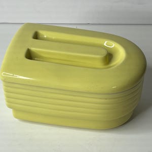 May include: A yellow ceramic trinket box with a lid. The box has a rectangular shape with rounded edges and a handle on top. The sides of the box are ribbed. The lid has a recessed handle. The box is a solid, pale yellow color.