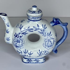 May include: White and blue ceramic teapot with a unique donut shape. The teapot features blue floral and leaf patterns, a spout, a handle, and a lid. The handle has a dragon-like design. A decorative kitchenware item.