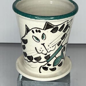May include: A white ceramic planter with a green rim features a hand-painted illustration of a cat with black spots holding a green fish. The planter has a matching saucer.
